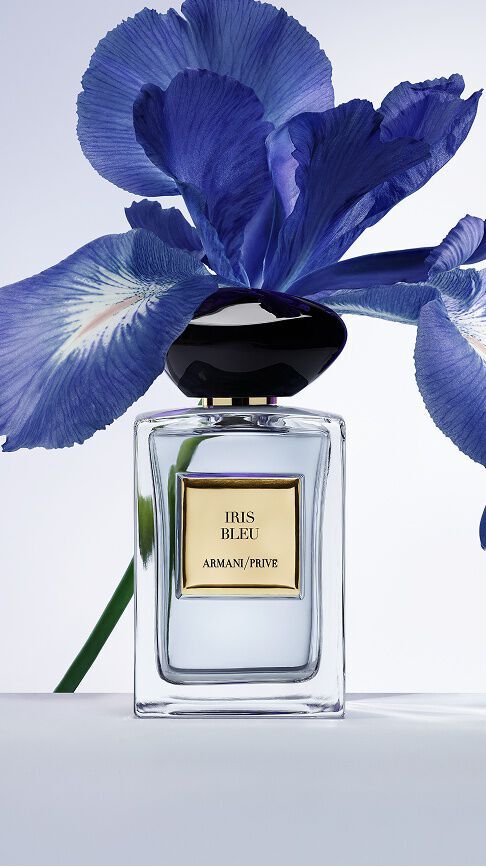 Armani/Prive Iris Bleu: een heldere glazen flacon met een zwarte stenen dop, geplaatst voor een levendige blauwe irisbloem, die een delicaat evenwicht tussen florale elegantie en diepte vangt.
