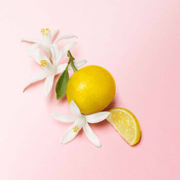 A vibrant yellow lemon with a slice beside it, accompanied by white blossoms and green leaves on a soft pink background.
