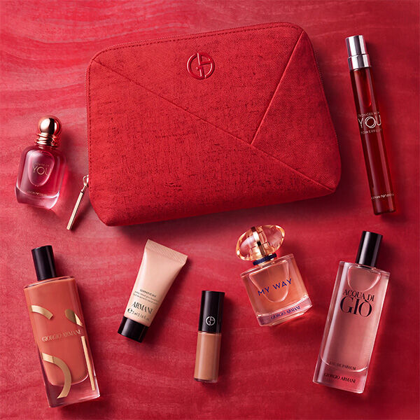 Een Armani zomercadeauset gepresenteerd op een wit marmeren rand met uitzicht op een rustige blauwe zee. In het midden staat een beige gebreide toilettas met een blauw geborduurd GA-logo. Links staan Si Passione, Acqua di Gio, een Luminous Silk foundation-sample, een vloeibare blush en een nude lipstick. Rechts staan de parfumminiaturen Armani Code, My Way en Power of You.