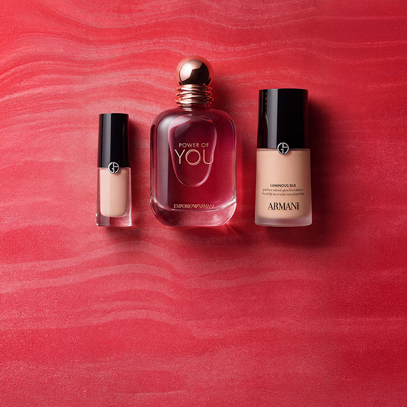 Een rode Armani Beauty-pouch, omringd door parfum-, make-up- en huidverzorgingsproducten tegen een rode achtergrond met textuur.