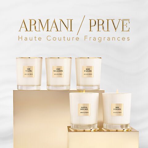 A display of five elegant candles in glass holders with gold accents, labeled with various names, featuring a soft white background.