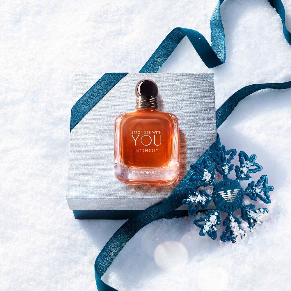 Armani beauty gift box — silver gift box wrapped with blue satin ribbon and a metallic snowflake ornament, resting on fresh white snow.
