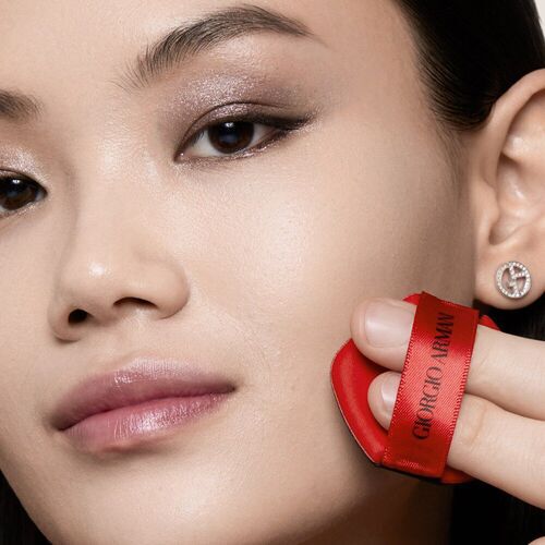 A close-up of a woman applying makeup with a red applicator sponge, featuring a subtle smoky eye and a natural glow on her skin. She is wearing a small earring and has a serene expression.