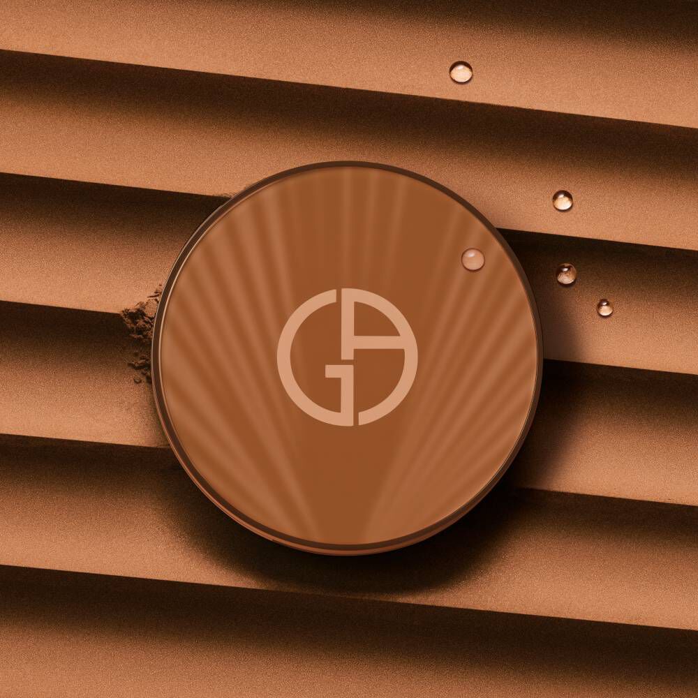 A round compact with a logo on its surface, placed on textured brown fabric, with a few water droplets nearby.