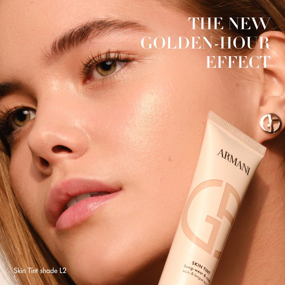 Vrouw met een stralende huid die een tube Armani Skin Tint vasthoudt, met de tekst THE NEW GOLDEN-HOUR EFFECT.
