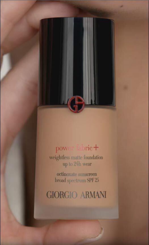 A hand holds a bottle of Giorgio Armani Power Fabric+ weightless matte foundation, featuring a black cap with a red logo and displaying details about its long-lasting wear and SPF 25 protection.