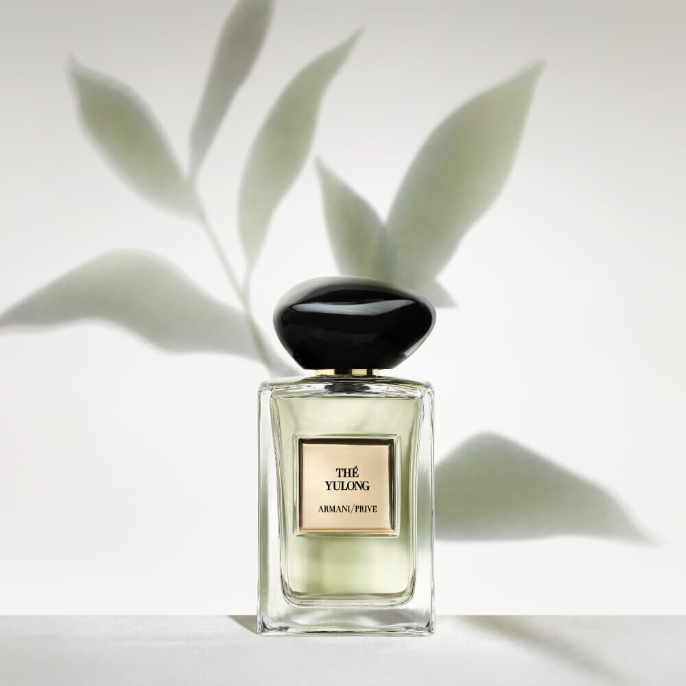Armani Prive Les Eaux The Yulong eau de parfum, transparent rectangular glass bottle with black rounded cap, set against delicate green botanical leaves on a pale sage background.