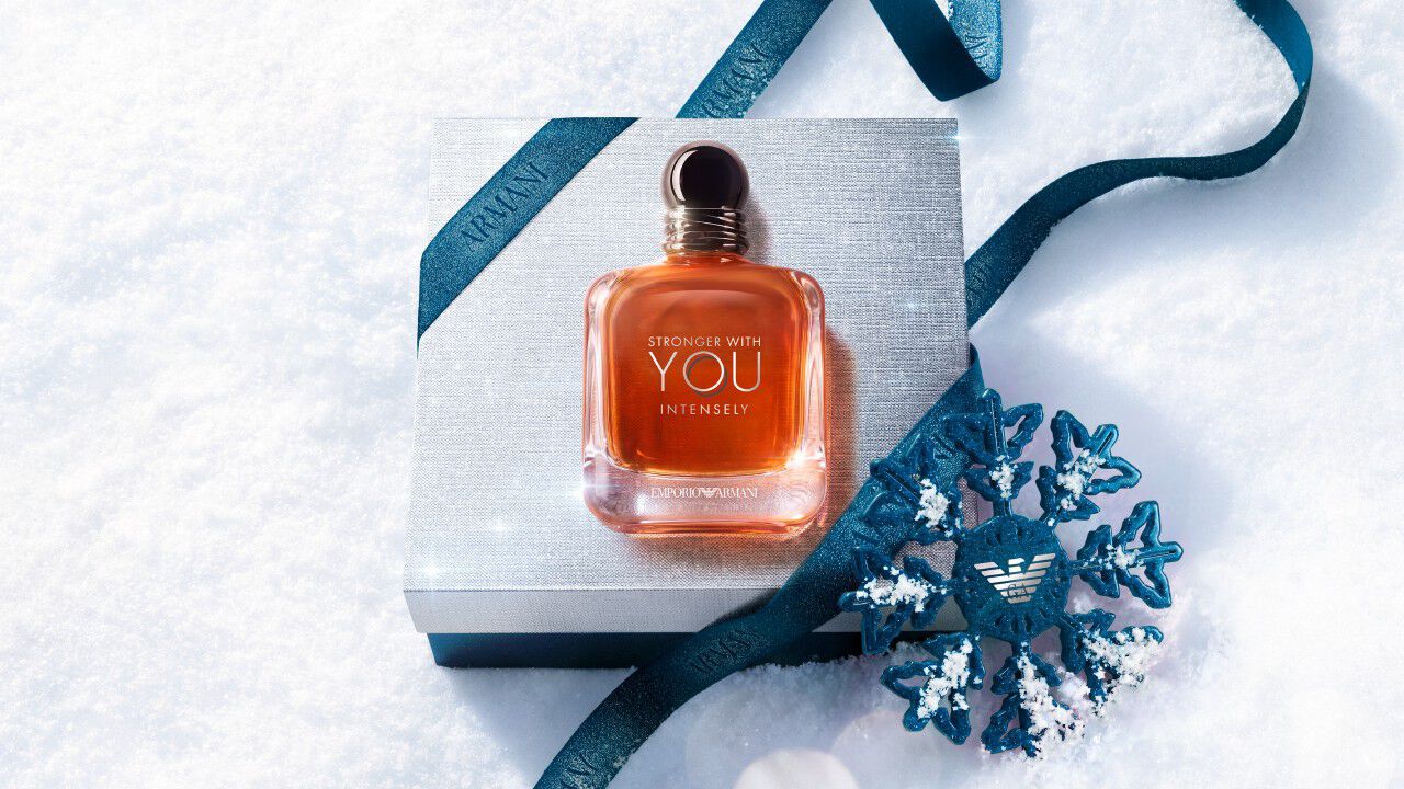 Armani beauty gift box — silver gift box wrapped with blue satin ribbon and a metallic snowflake ornament, resting on fresh white snow.