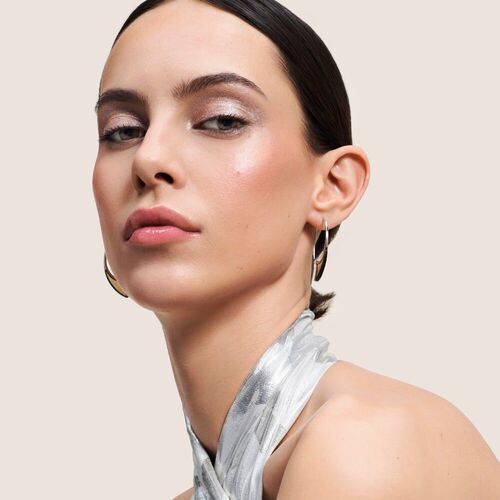 Model met een strakke updo, glanzende huid en zilveren halsketting, poseert op een neutrale achtergrond.
