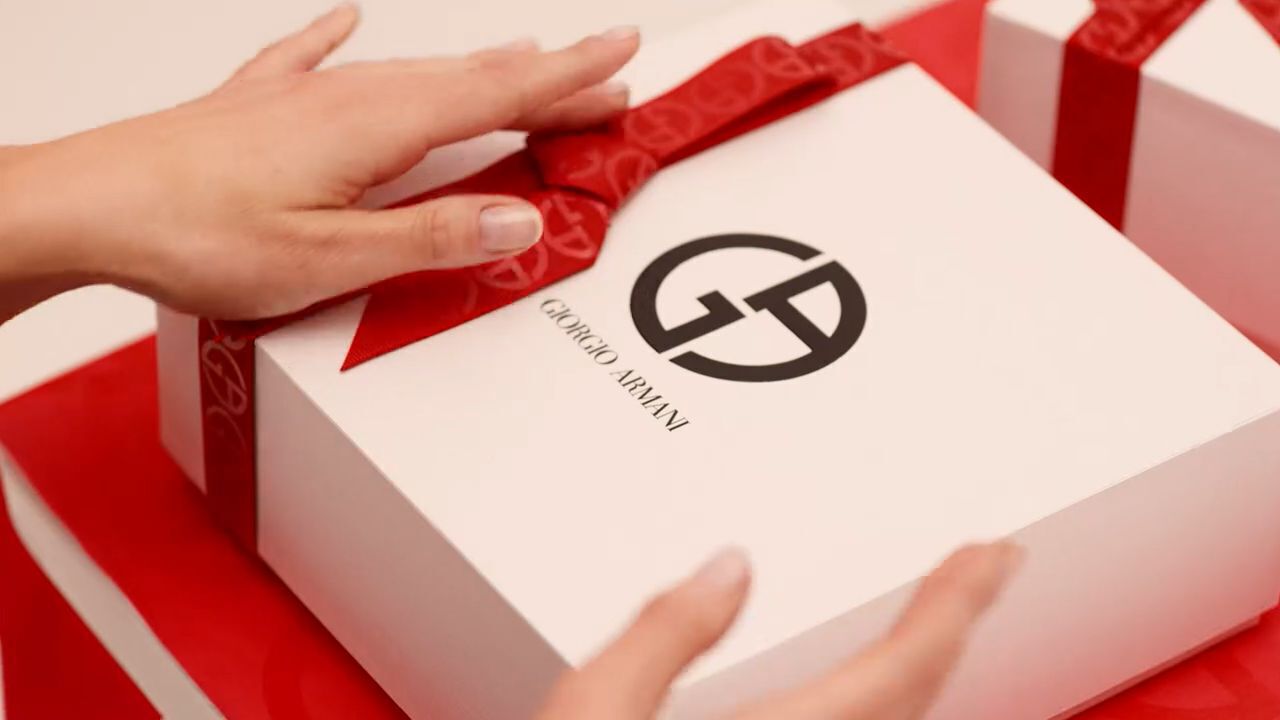 A close-up of a hands delicately placing a ribbon on a white gift box featuring the Giorgio Armani logo, set against a background of red wrapping.