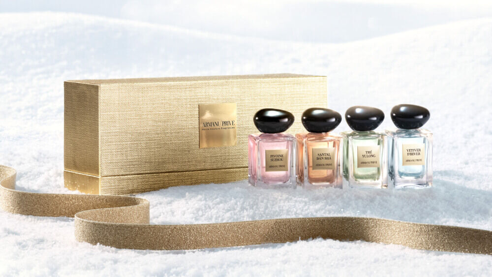 Armani Prive Les Eaux Holiday miniature gift set: gold rectangular box alongside four miniature eau de parfum bottles, photographed on a pale textured snow surface.
