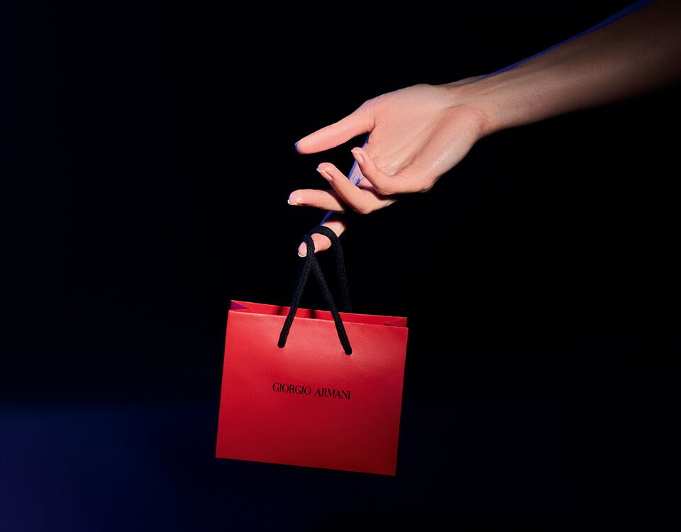 A hand holds a small red shopping bag with black handles, featuring the name "Giorgio Armani" in black text. The background is dark, emphasizing the bag.