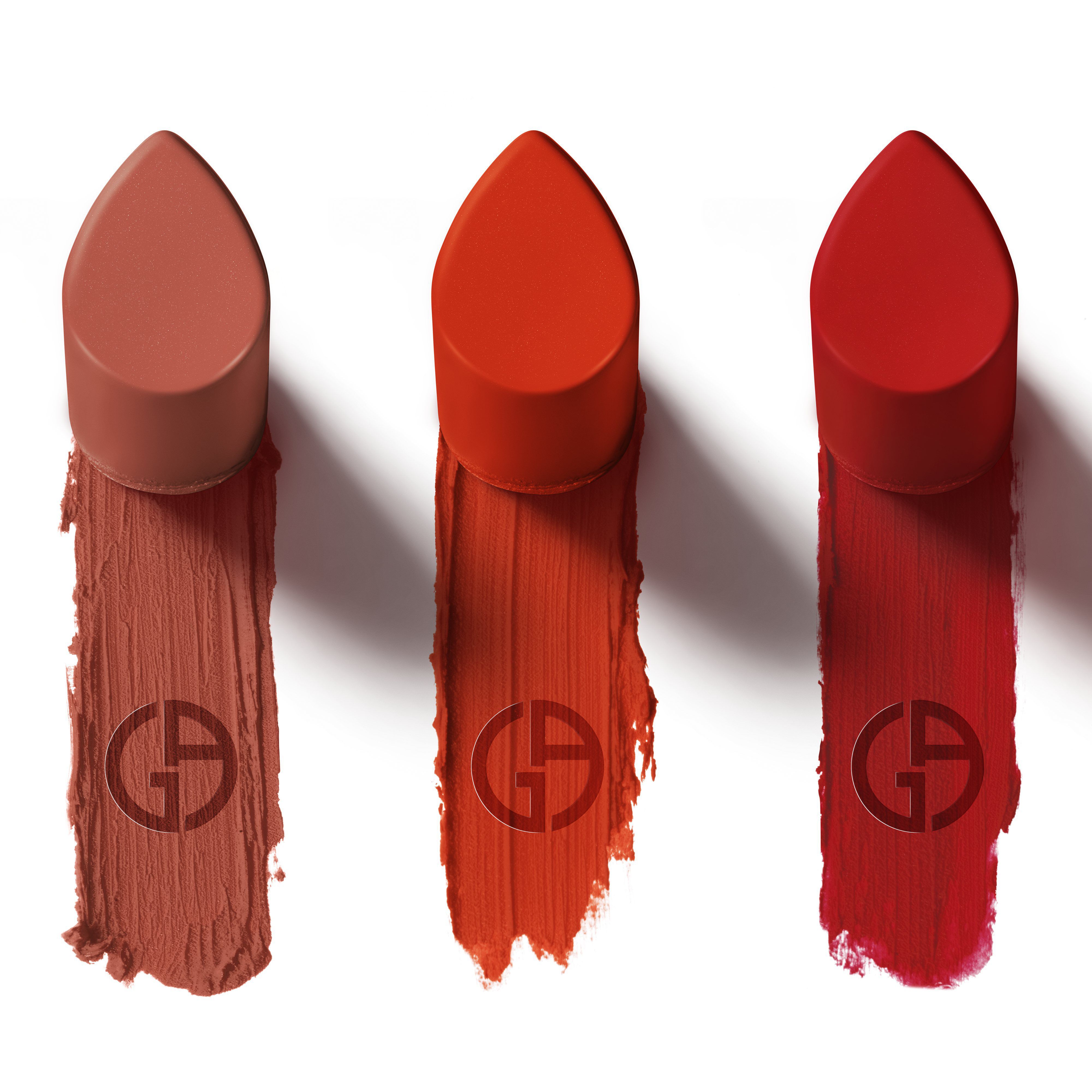 Three lipsticks are displayed in a row, each with a color-matched cap. Below them are streaks of lipstick in corresponding shades: a nude tone on the left, a vibrant orange-red in the middle, and a bold red on the right. All have the logo of the brand "Giorgio Armani" embossed in the swatches.