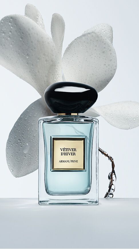 Armani/Prive Vetiver d'Hiver: een glazen flacon met lichtblauw parfum, omringd door zachte witte bloembladen met dauwdruppels, die de kristalheldere en stralende frisheid van de winter oproept.