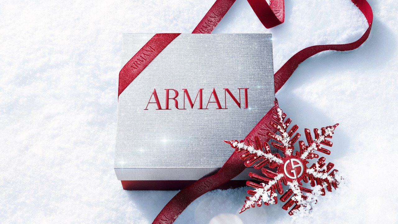 Silver Armani Glamourous Winter Holiday Gift Set box with red glitter ribbon and Giorgio Armani red snowflake ornament resting on a sparkling snowy surface — festive luxury fragrance gift packaging.