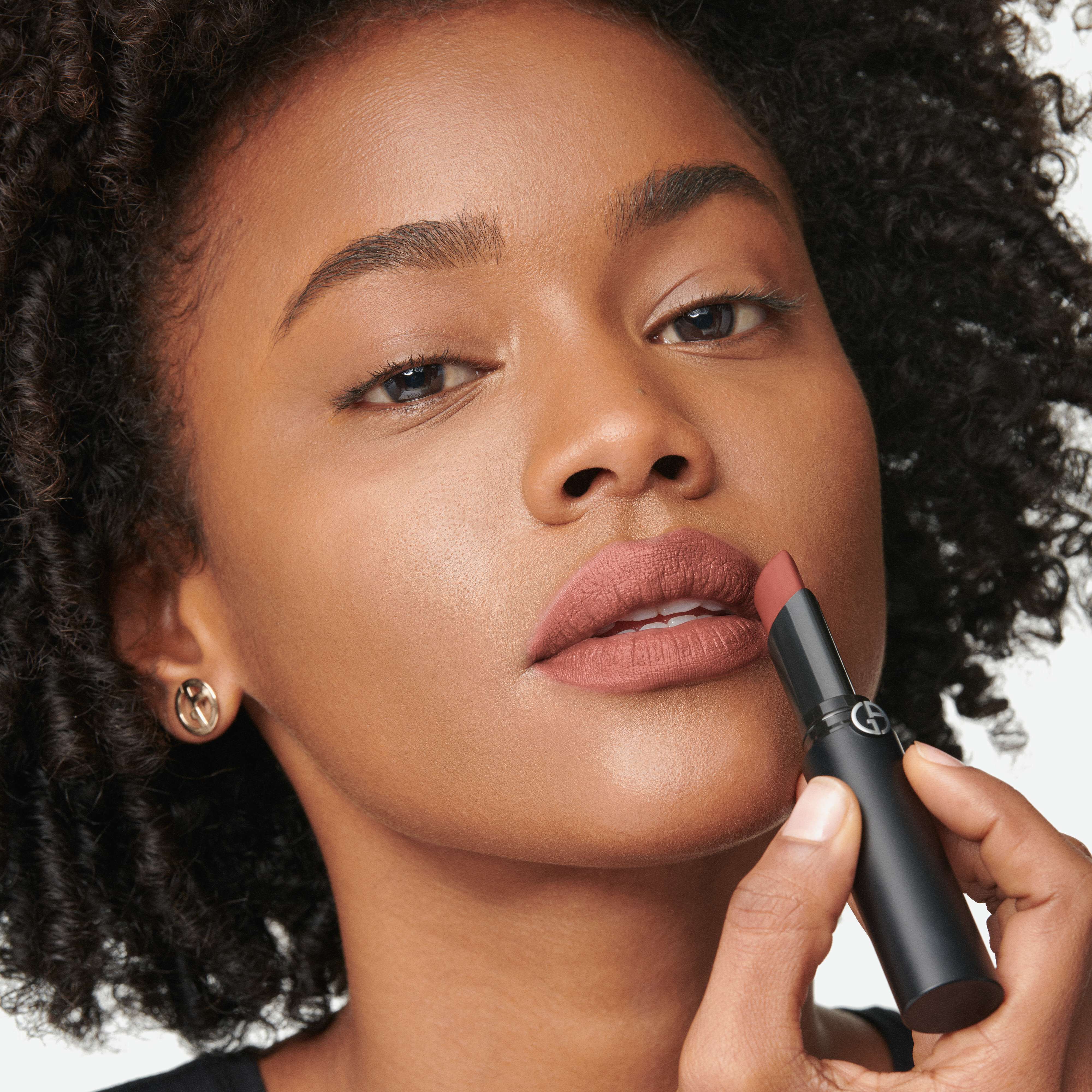 A close-up of a woman with curly hair applying a mauve lipstick, holding the lipstick tube close to her lips. She has a natural makeup look and is wearing small earrings.