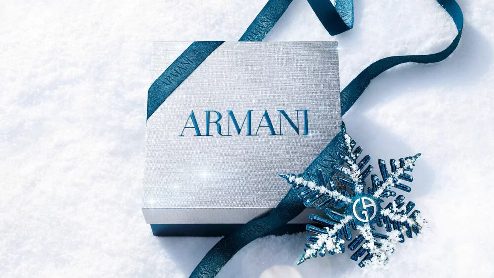 Armani beauty gift box — silver gift box wrapped with blue satin ribbon and a metallic snowflake ornament, resting on fresh white snow.