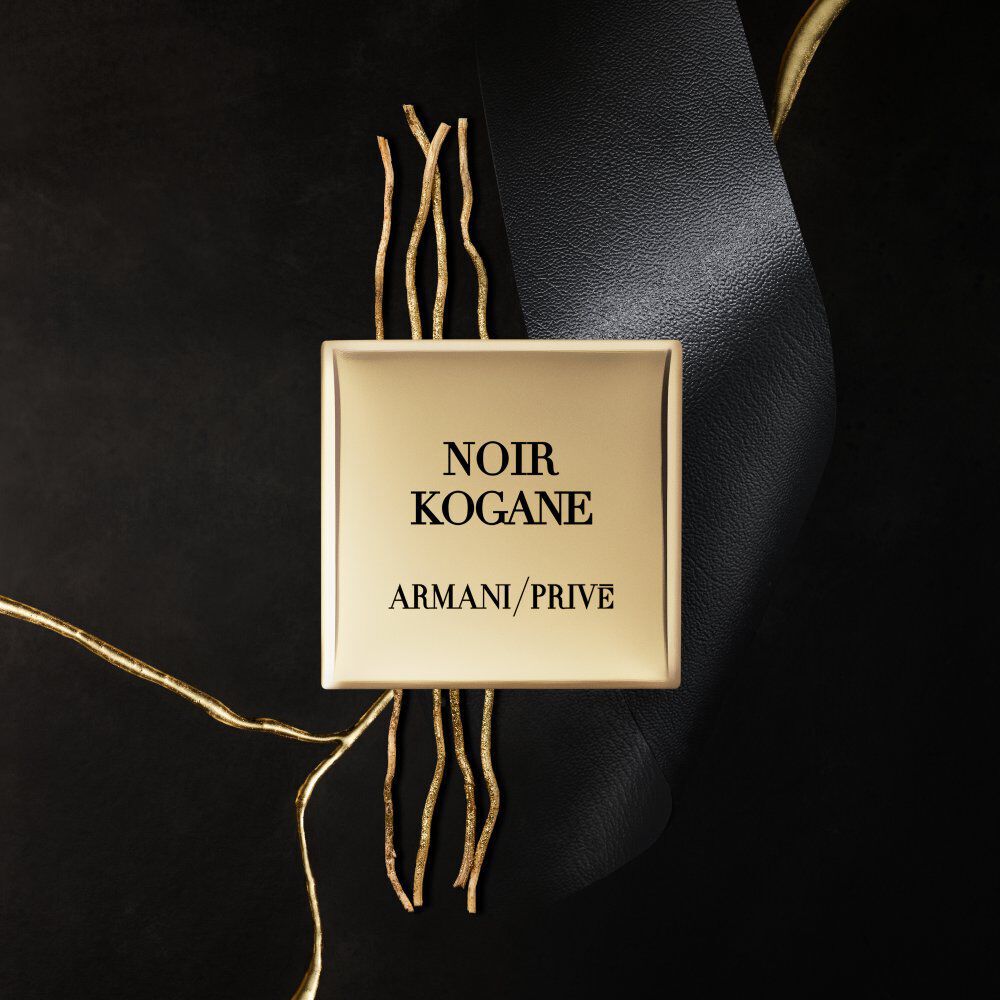 A gold rectangular box with the text "NOIR KOGANE" and "ARMANI/PRIVE" printed in black, surrounded by a black ribbon and thin golden twigs on a dark background.