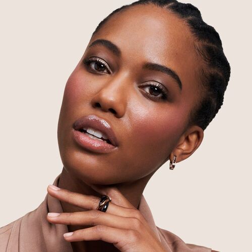 Model met een stralende huid, een elegante ring en oorbellen, kijkt recht in de camera tegen een lichte achtergrond.