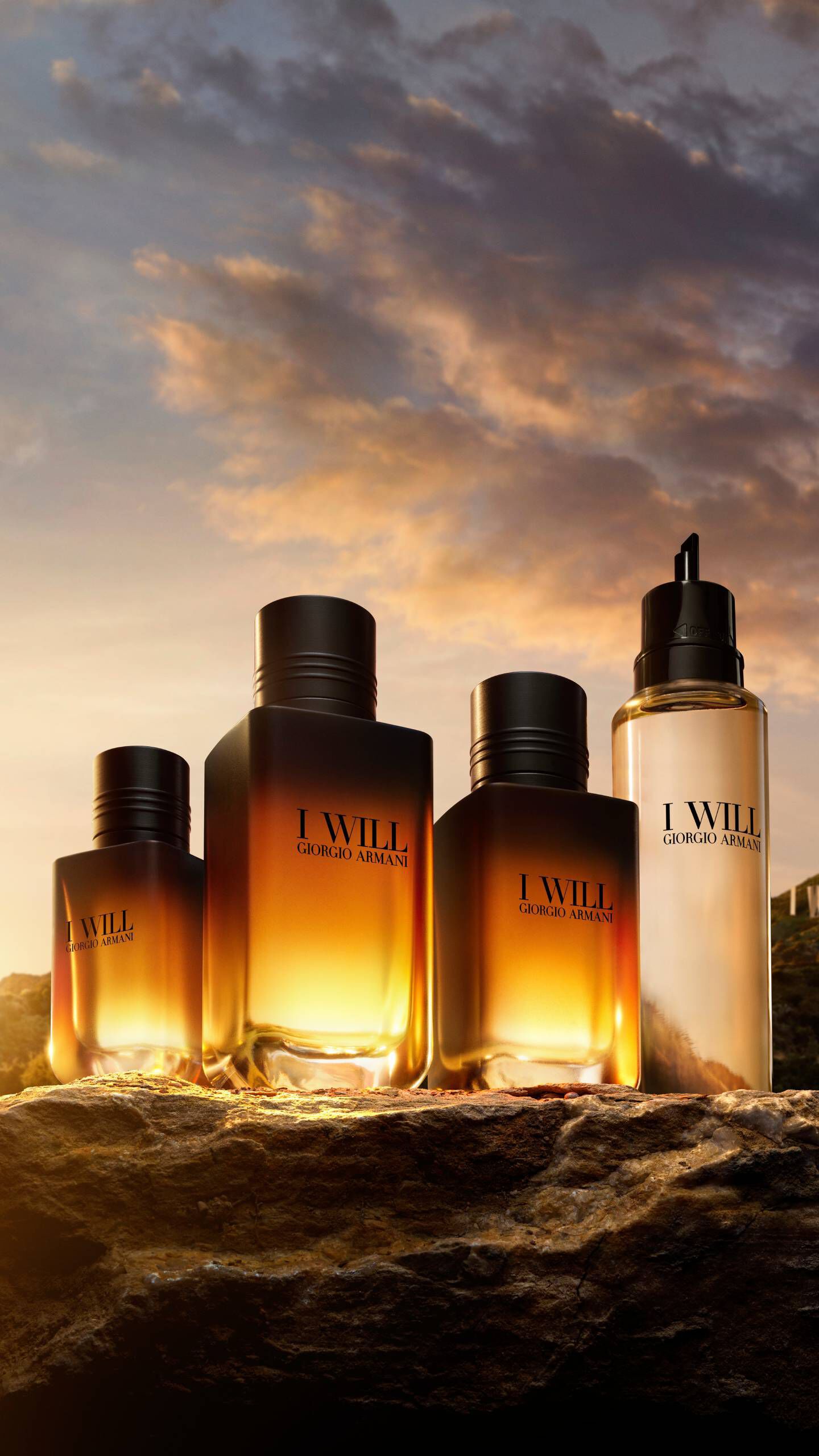 Flacons van Giorgio Armani I WILL Eau de Parfum in verschillende formaten, gepresenteerd op een rotsachtig landschap tijdens het gouden uur, waarbij het warme licht van de ondergaande zon door de amberkleurige geurvloeistof gloeit.