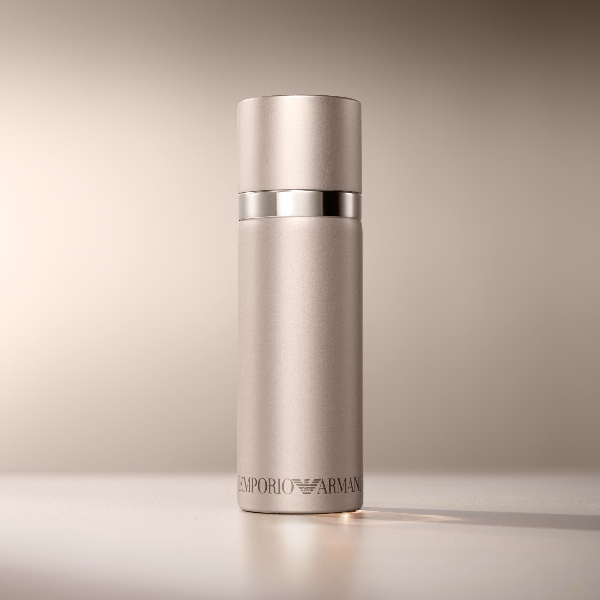 Een matte rozencilindrische Emporio Armani "She" parfumfles is elegant geplaatst op een warme, lichtbruine achtergrond.