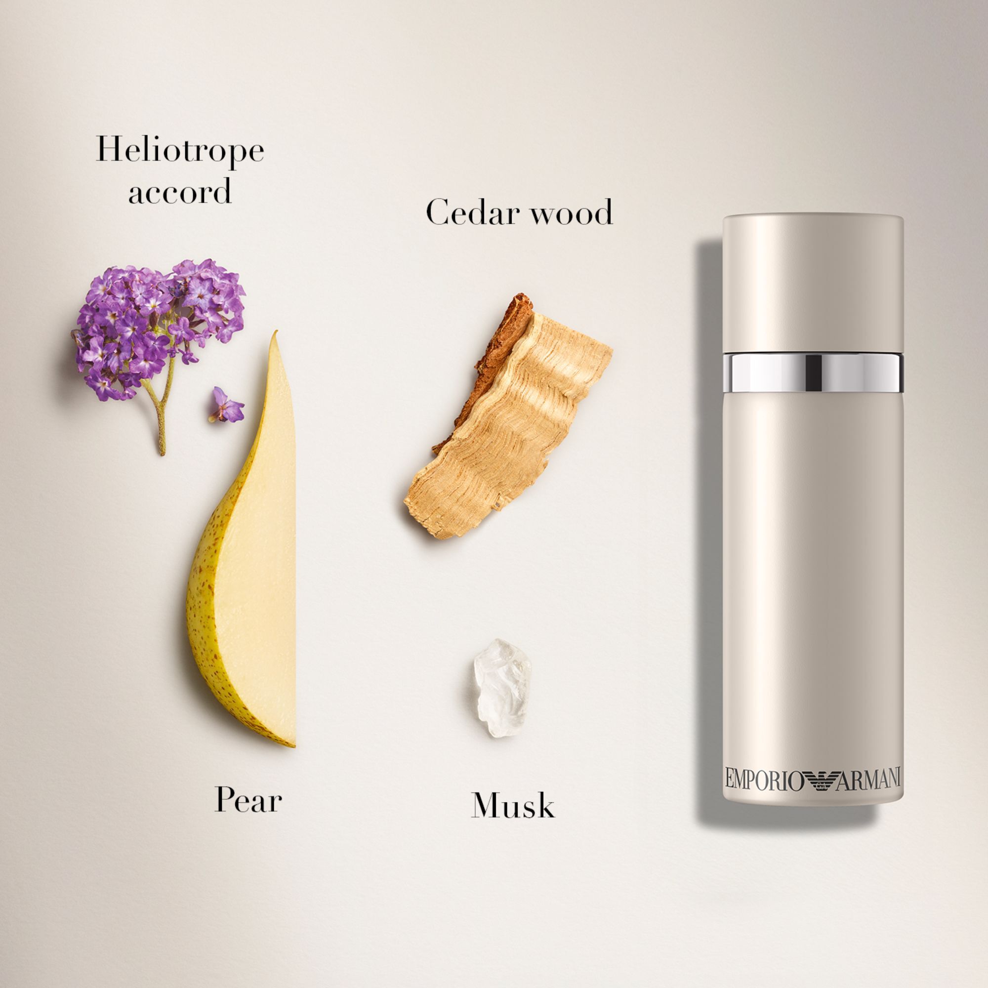 Een platte lay-compositie voor de Emporio Armani "She" geur, met een 100ml lichtbeige fles naast ingredienten zoals heliotrope accord (paarse bloemen), cederhout, een gesneden peer en muskus, op een lichte achtergrond.
