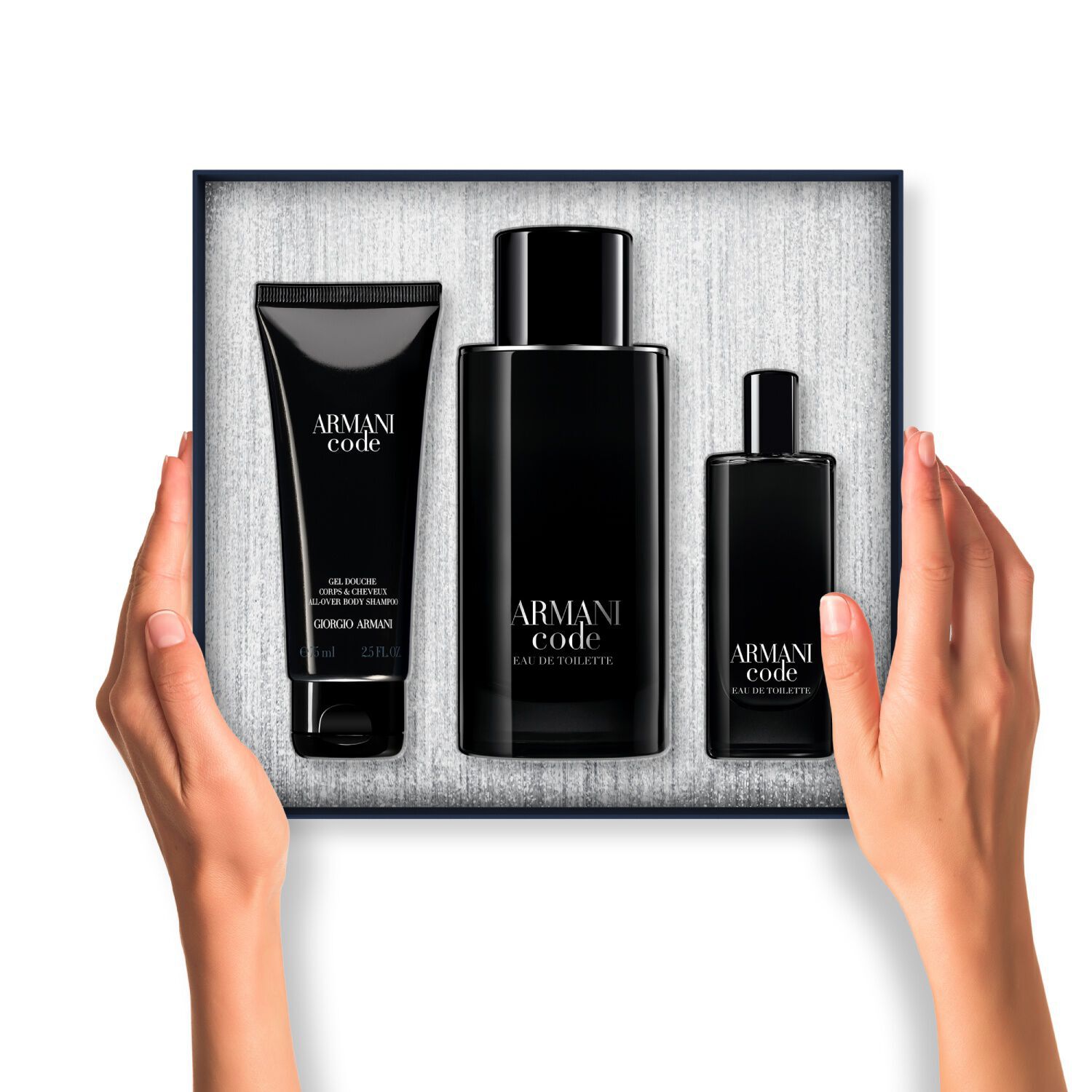 Een paar handen houdt een open Armani Code cadeauverpakking vast. Binnenin zijn drie zwart verpakte producten (bodygel, grote Eau de Toilette en reisformaat Eau de Toilette) netjes gerangschikt tegen een getextureerde zilveren achtergrond.