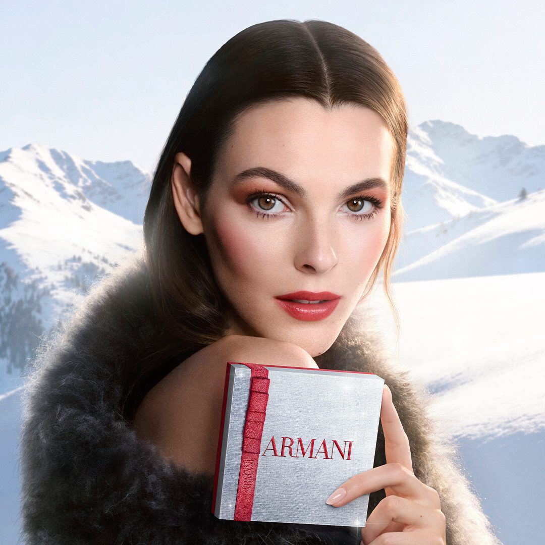 Campaign key visual: Vittoria Ceretti in a winter fur coat holding an Armani beauty Holiday 2026 white gift box, set against a snowy mountain backdrop.