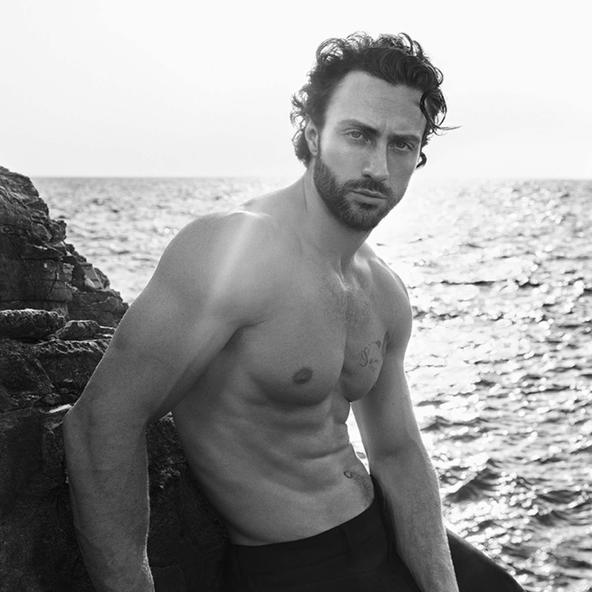 Aaron Taylor-Johnson aan zee, tegen een rotsachtige kustlijn. Een zwart-witte mariene atmosfeer.