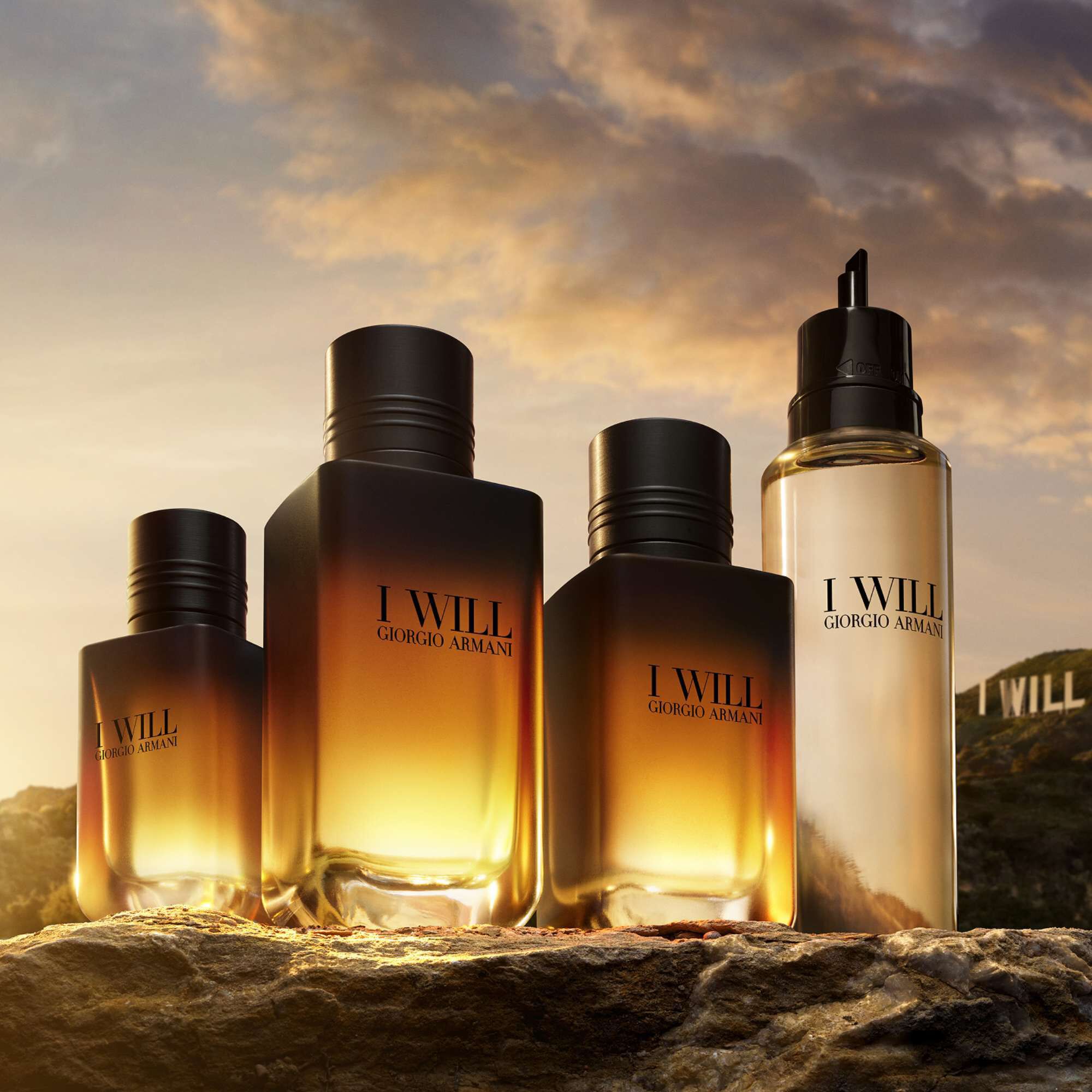 Flacons van Giorgio Armani I WILL Eau de Parfum in verschillende formaten, gepresenteerd op een rotsachtig landschap tijdens het gouden uur, waarbij het warme licht van de ondergaande zon door de amberkleurige geurvloeistof gloeit.