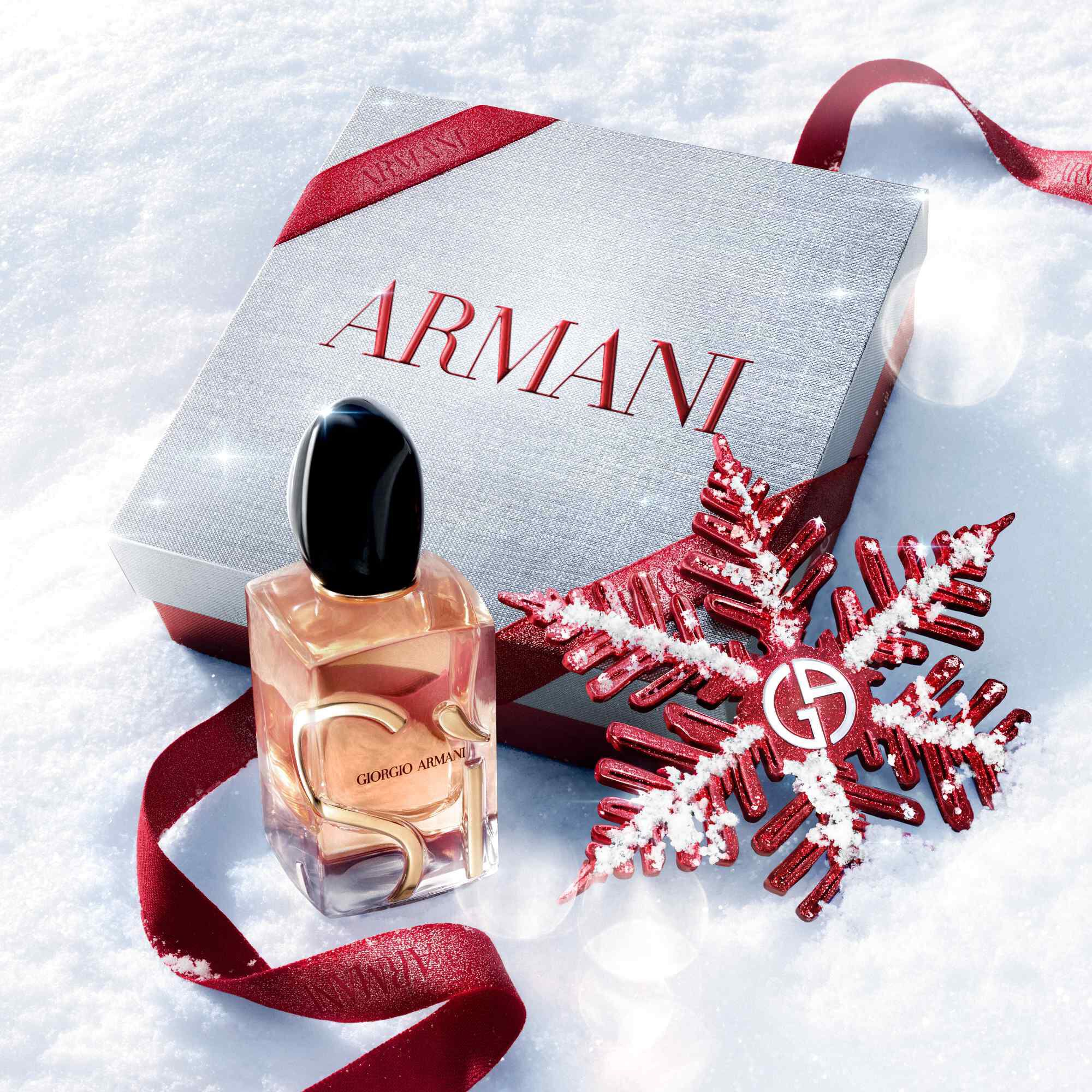 Si Eau de Parfum van Giorgio Armani, goudkleurige fles rustend in de sneeuw met een rood glitterlint en Giorgio Armani sneeuwvlokornament — damesgeurfeest