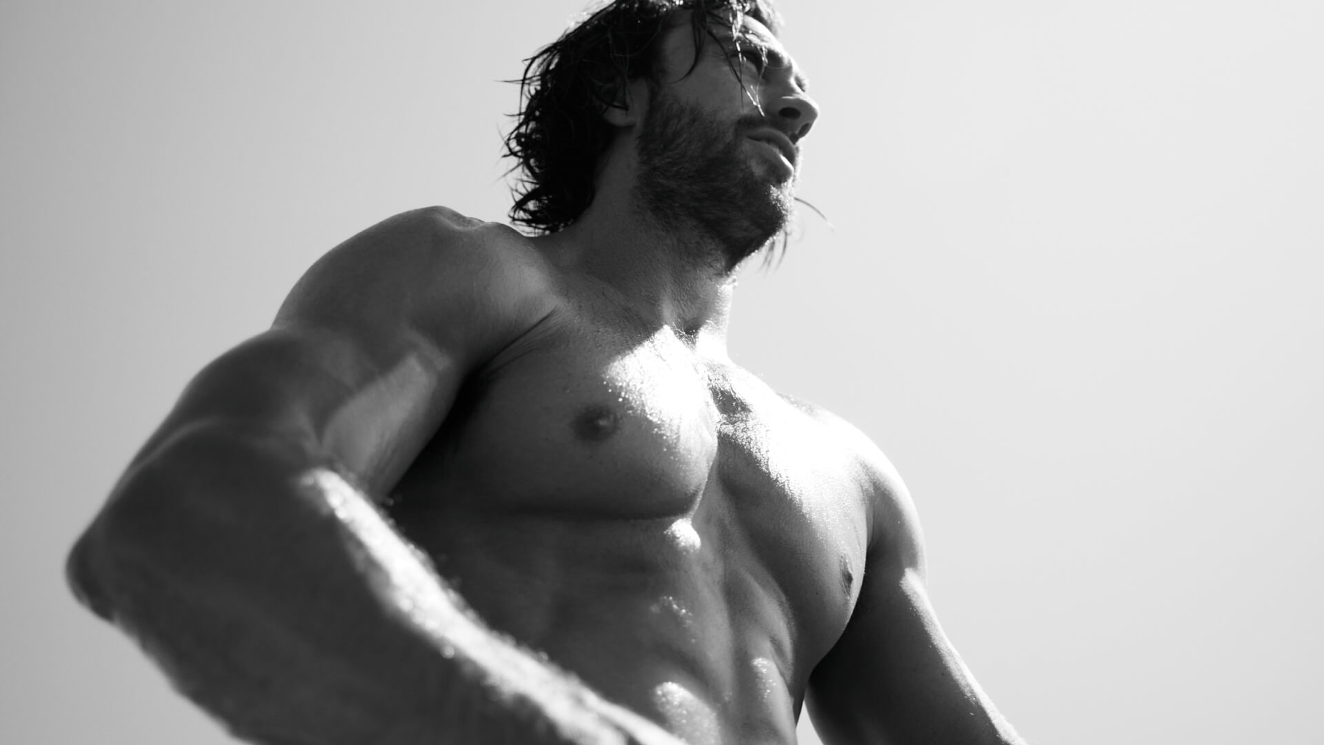 Een gespierde man zonder shirt staat in het zonlicht met een intense blik, gefotografeerd vanuit een laag perspectief.