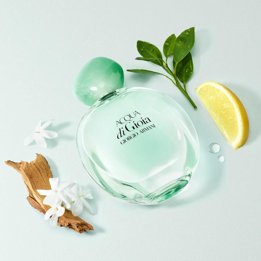 Een flesje Acqua di Gioia parfum met groene dop, omringd door citroen, bloesems en bladeren op een zachte achtergrond.