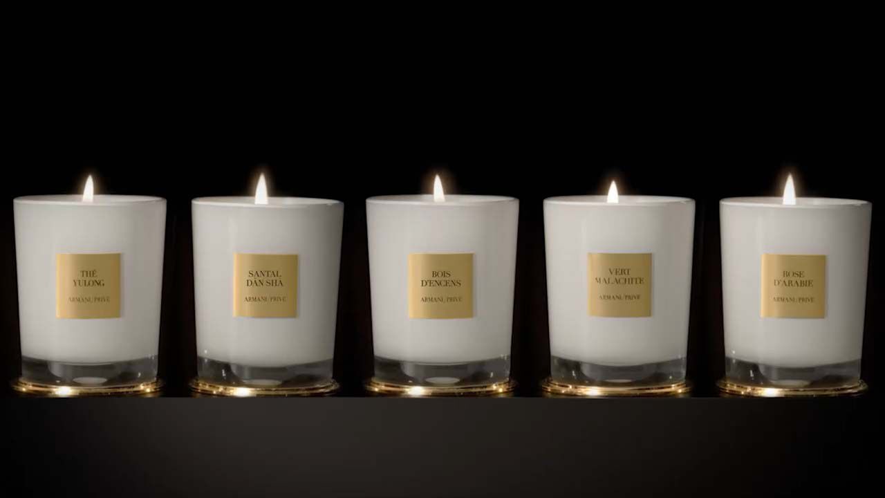 Vijf brandende kaarsen in witte glazen met gouden etiketten, elk een unieke geur van aromatische producten.